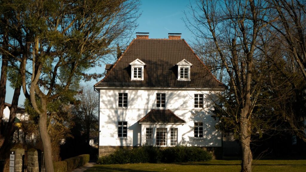 pexels-photo-31444161-1 Elegant colonial-style house with traditional roof amid lush trees on a sunny day.
